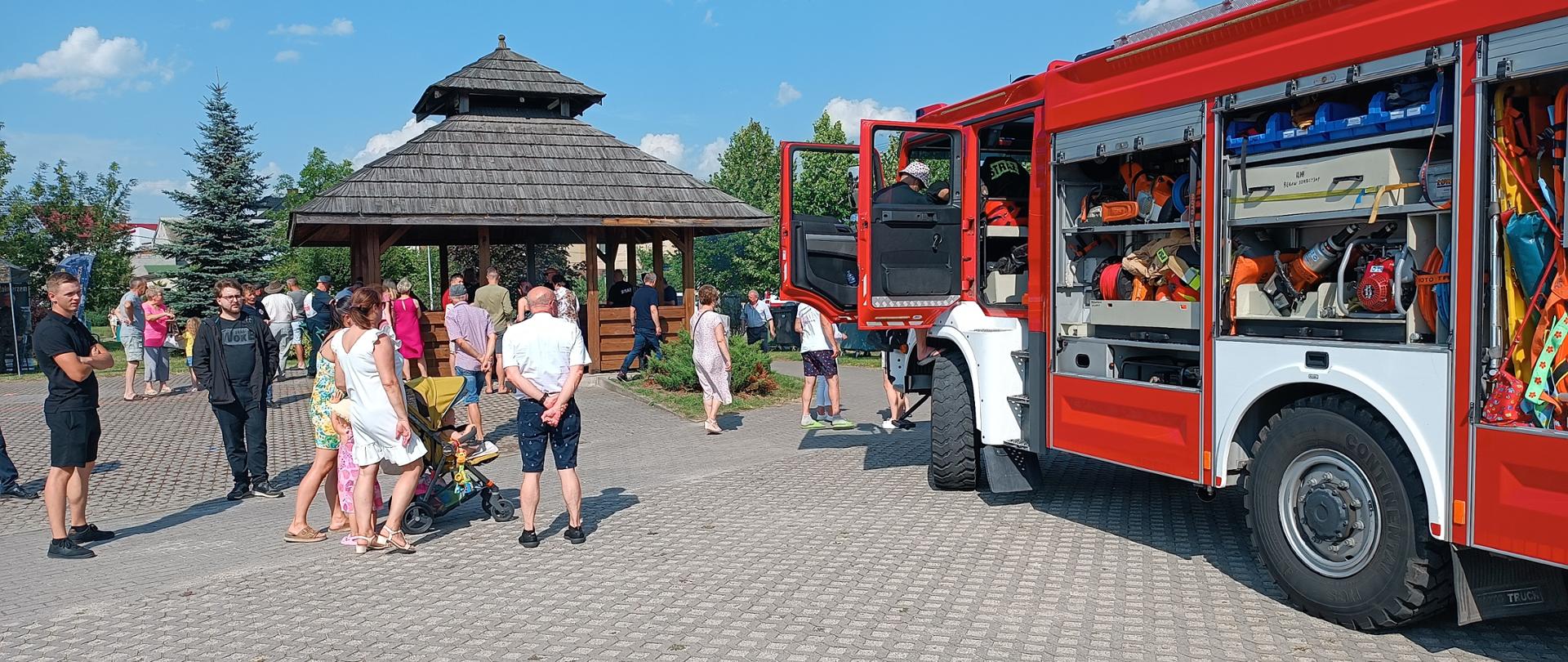 Po prawej stronie samochód strażacki, na prosto ludzie . w tle altanka, 