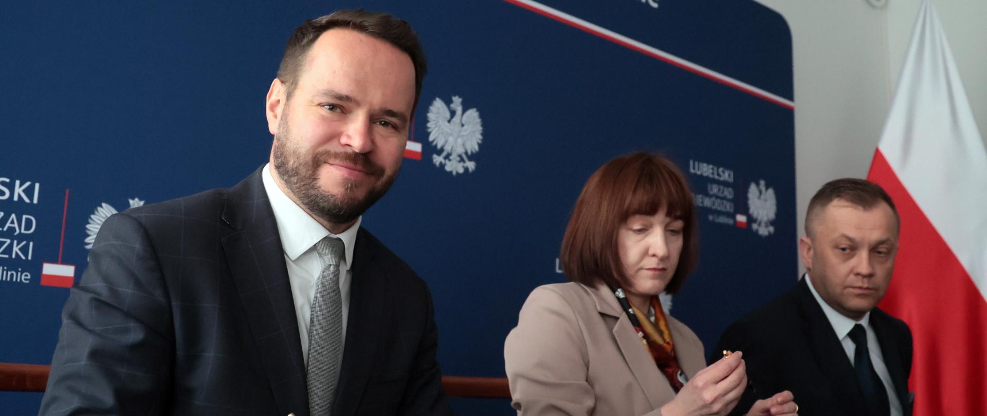 Uśmiechnięty mężczyzna w garniturze podpisuje dokument przy stole. Obok siedzą kobieta przyglądająca się złotemu przedmiotowi, najprawdopodobniej długopisowi i poważnie wyglądający mężczyzna. W tle widnieje baner "Lublinie" z godłem Polski i fragment polskiej flagi.