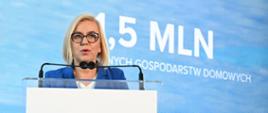 Osoba w niebieskiej marynarce stoi za przezroczystą mównicą i przemawia do mikrofonów podczas konferencji. W tle widoczny jest niebieski ekran z dużym napisem „1,5 MLN” oraz fragmentem tekstu odnoszącego się do gospodarstw domowych. Zdjęcie wykonano we wnętrzu, w trakcie wystąpienia publicznego.