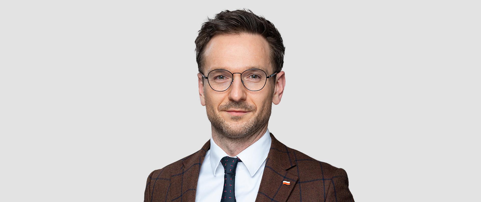 Minister rozwoju i technologii Waldemar Buda, minister ubrany jest w białą koszulę z granatowym krawatem i brązową marynarkę w klapę której ma wpiętą znaczek z polską flagą 