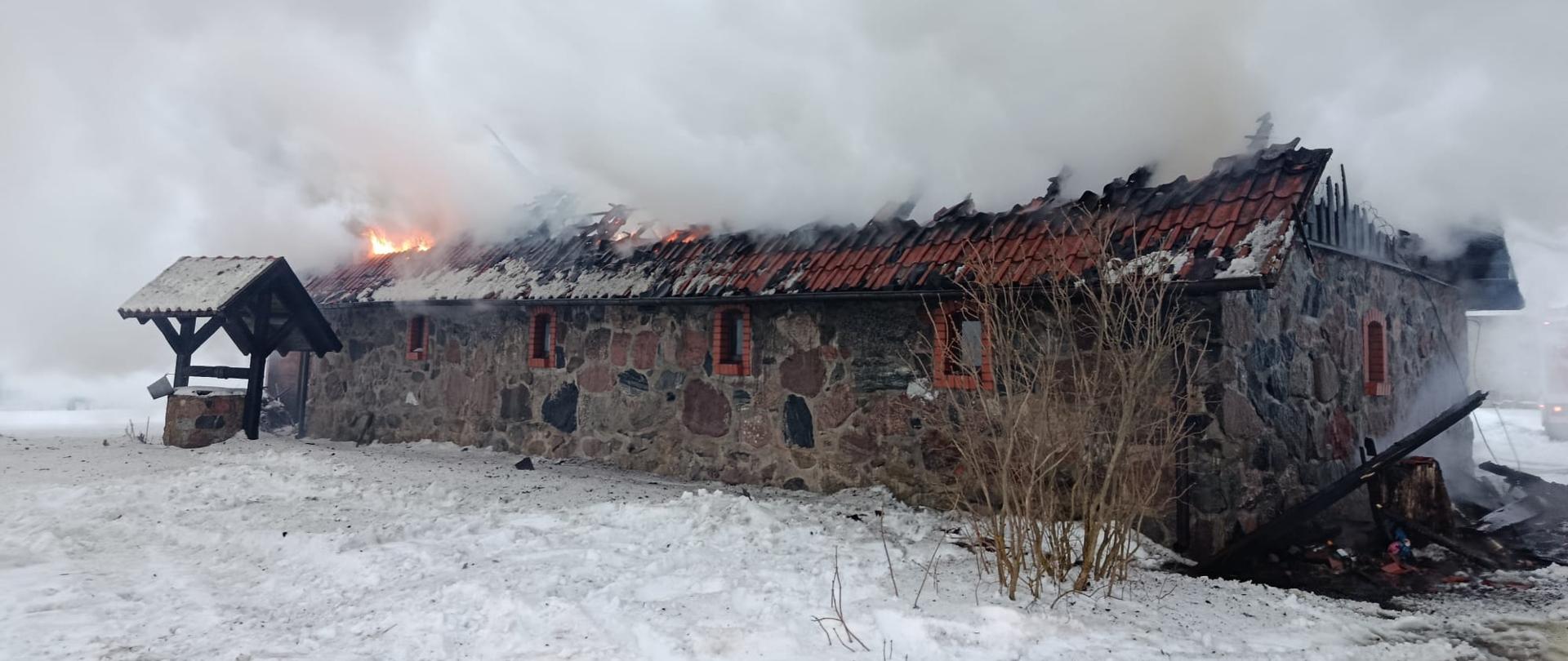 Pożar murowanego budynku gospodarczego – widoczny częściowo zawalony dach objęty ogniem i kłęby dymu unoszące się nad obiektem; teren pokryty śniegiem, w tle działania straży pożarnej.