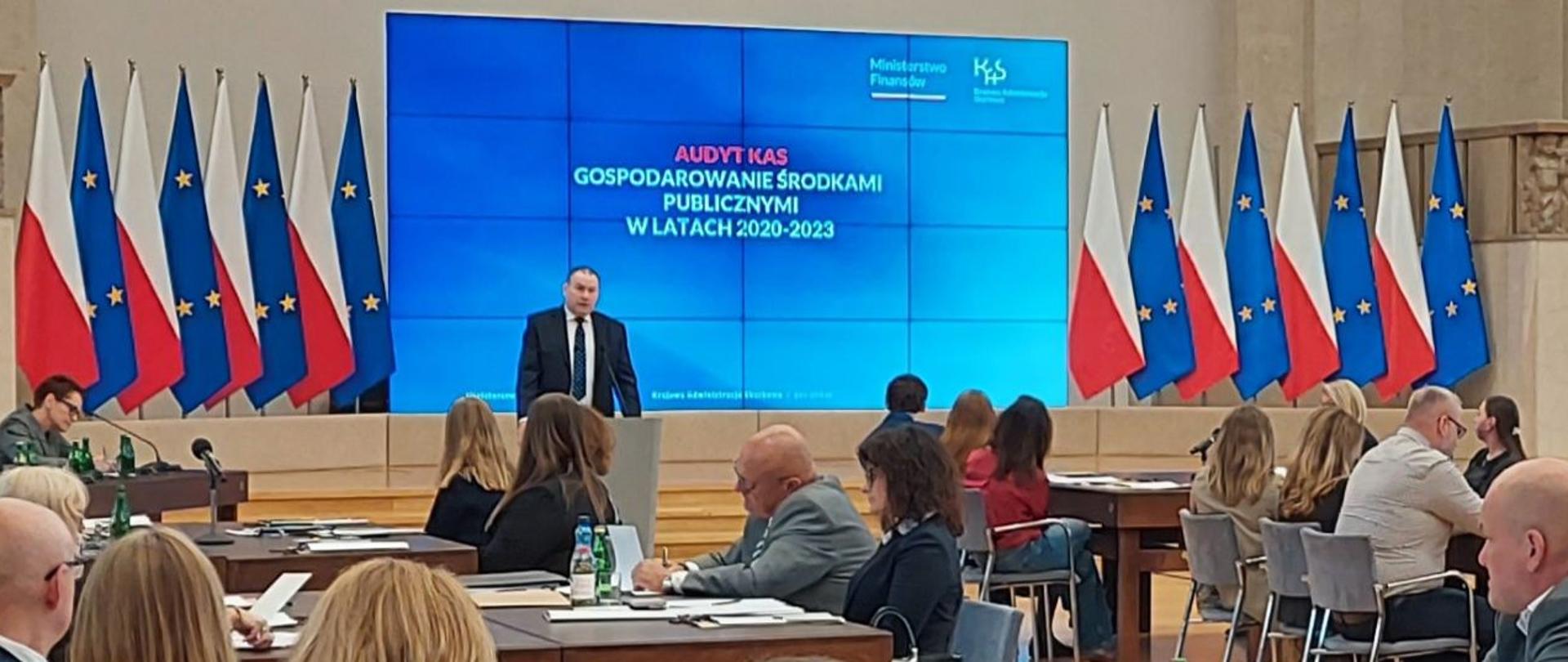 W dużej sali konferencyjnej uczestnicy siedzą przy stołach i słuchają z-cy Szefa KAS Zbigniewa Stawickiego przy mównicy; w tle widoczny jest duży ekran z prezentacją oraz rzędy flag Polski i Unii Europejskiej.
