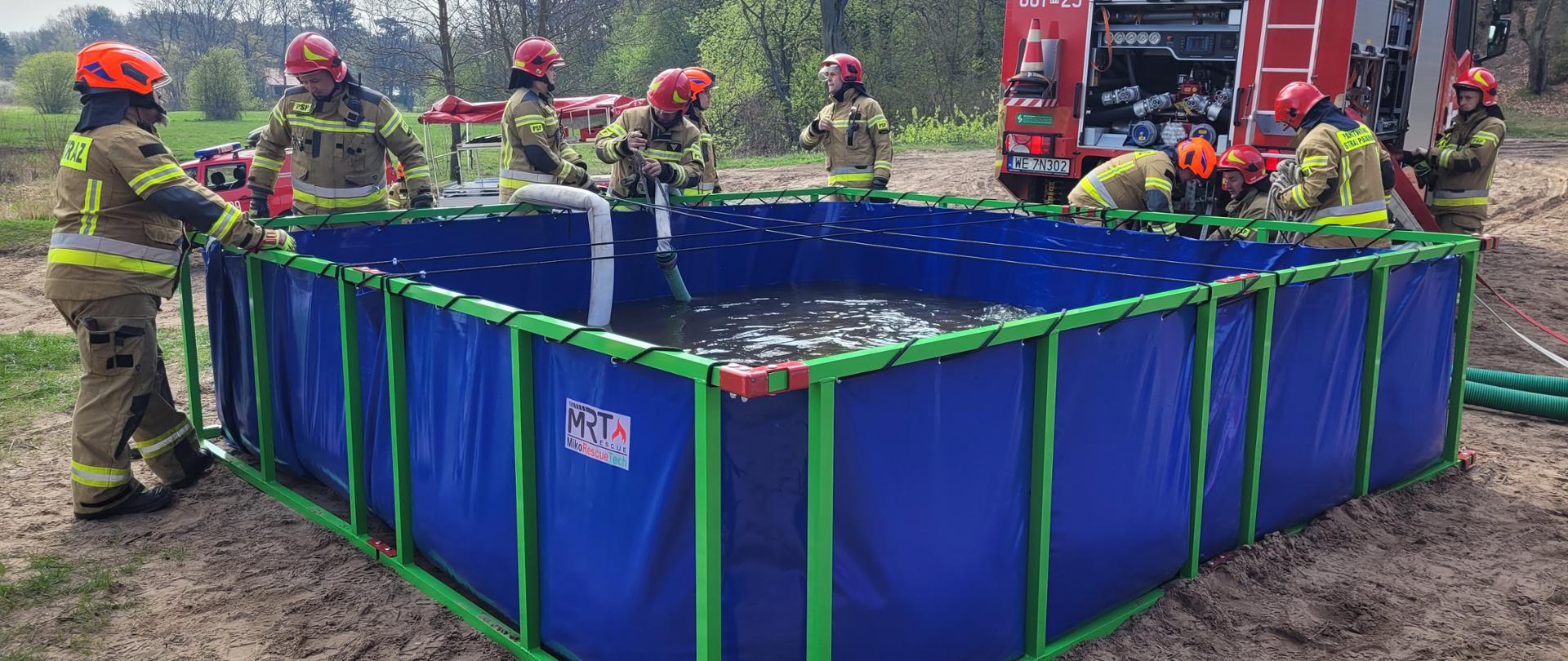 Fotografia przedstawia strażaków stojących przy składanym niebieskim zbiorniku na wodę. Zbiornik ustawiony jest nad brzegiem rzeki. Wokoło znajdują się strażackie samochody a w tle widać niebieskie niebo. Jeden ze strażaków schodzi z dachu pojazdu pożarniczego. 