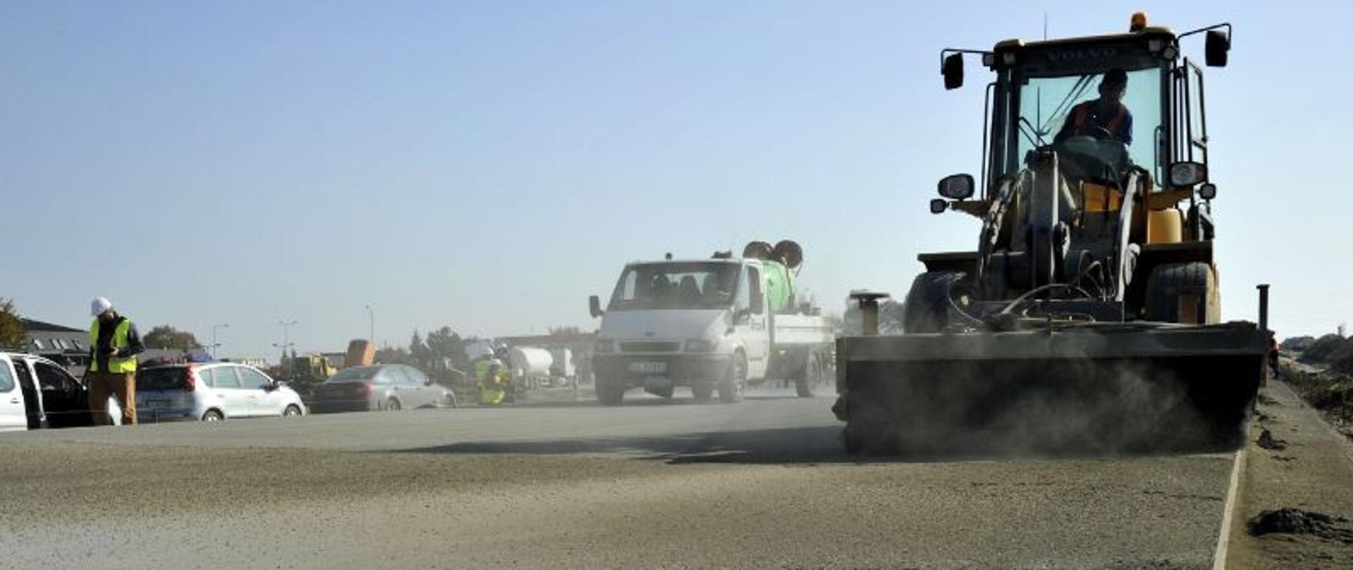 Plac budowy, z prawej maszyna układa nawierzchnię drogi , w tle pojazdy budowy 