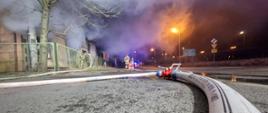 Widok z perspektywy - wąż strażacki z rozdzielaczem a w tle płonący budynek
