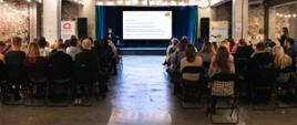 Tyłem do kamery w ośmiu rzędach siedzą uczestnicy spotkania i słuchają kobiety, która mówi do mikrofonu stojąc obok ekranu z wyświetlonym slajdem prezentacji