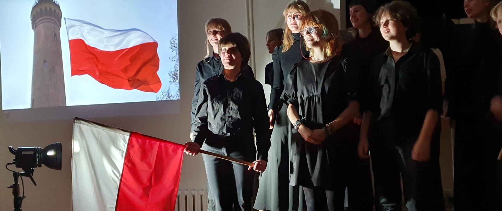 Zdjęcie - w centrum uczniowie, w czarnych strojach. Jedna postać trzyma flagę biało - czerwoną.