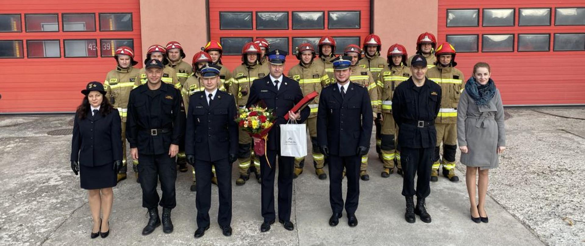 Na tle czerwonych trzech bram garażowych stoją strażacy w dwuszeregu w żółtych ubraniach bojowych. Przed nimi w rzędzie stoją strażacy w granatowych mundurach. Strażak stojący po środku trzyma upominki i kwiaty.