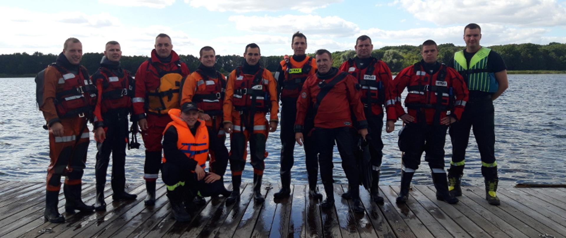 Szkolenia z ratownictwa na obszarach wodnych. Grupa strażaków ubrana w skafandry nurkowe i kamizelki ratunkowe stoi na pomoście i pozuje do wspólnego, pamiątkowego zdjęcia. W tle jezioro i drzewa.