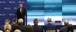
Dyrektor Departamentu Programów Wsparcia Innowacji i Rozwoju w MFiPR Grzegorz Orawiec stoi przy mównicy i przemawia do mikrofonu, za jego plecami telebim informujący o nowej formie finansowania firm
