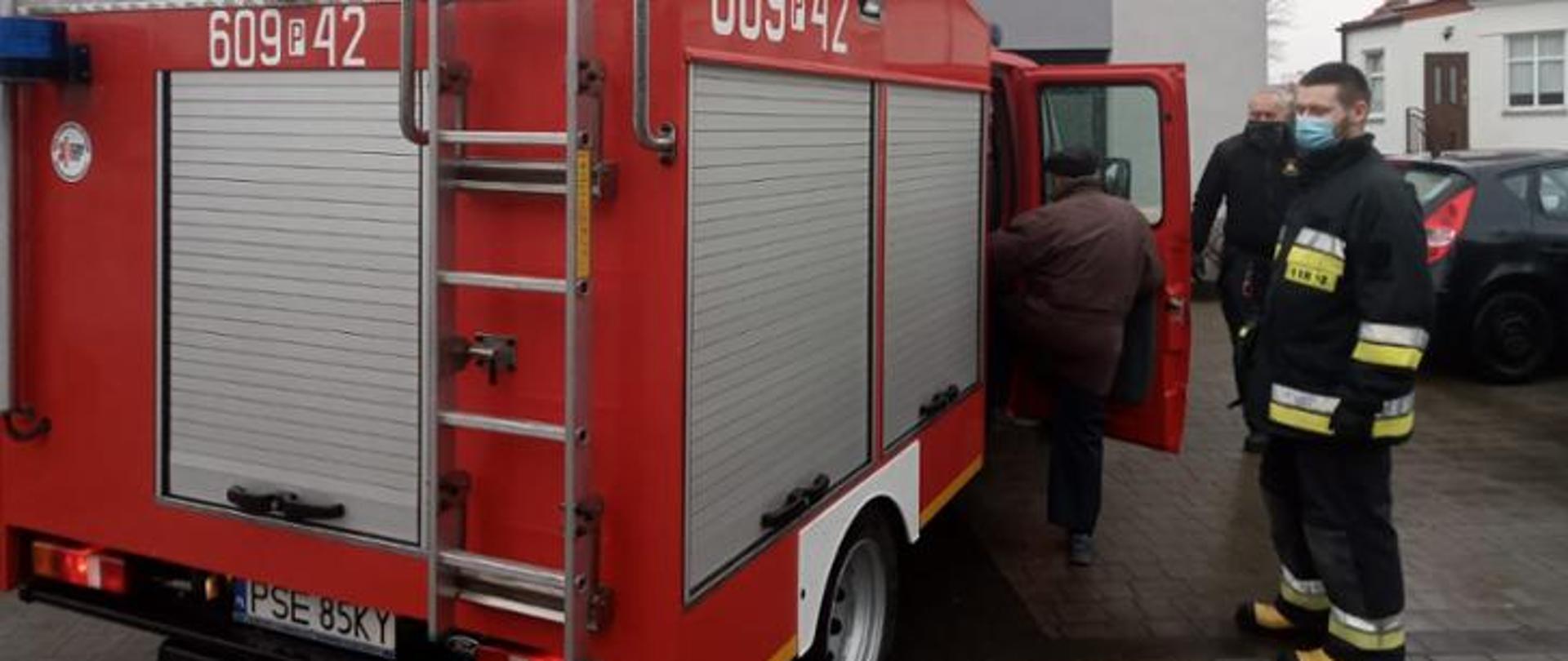 Pomoc w transporcie straszej osoby na szczepienie, mężczyzna wsiada do pojazdu pożarncizego w asyście strażaka i opiekuna