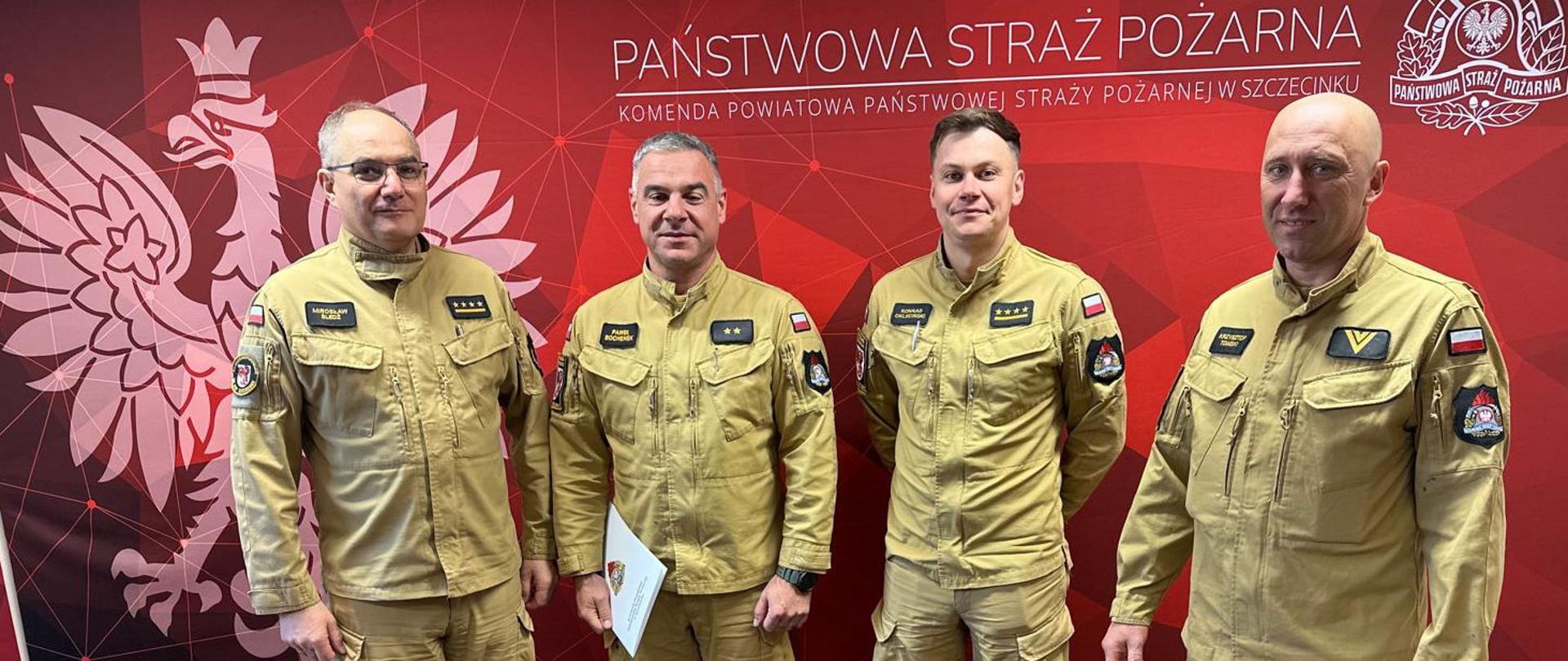 czterech mężczyzn w mundurach Państwowej Straży Pożarnej stoi w biurze. Po prawej stronie jeden z nich komendant powiatowy starszy kapitan Mirosław Śledź trzyma w rękach dokument, decyzję o nadaniu nagrody jubileuszowej i czyta ją, zwracając się do drugiego strażaka, który stoi naprzeciwko niego w postawie zasadniczej. W tle znajduje się ścianka z logo PSP Komendy Powiatowej Państwowej Straży Pożarnej w Szczecinku. Scena jest podczas wręczenia nagrody jubileuszowej.