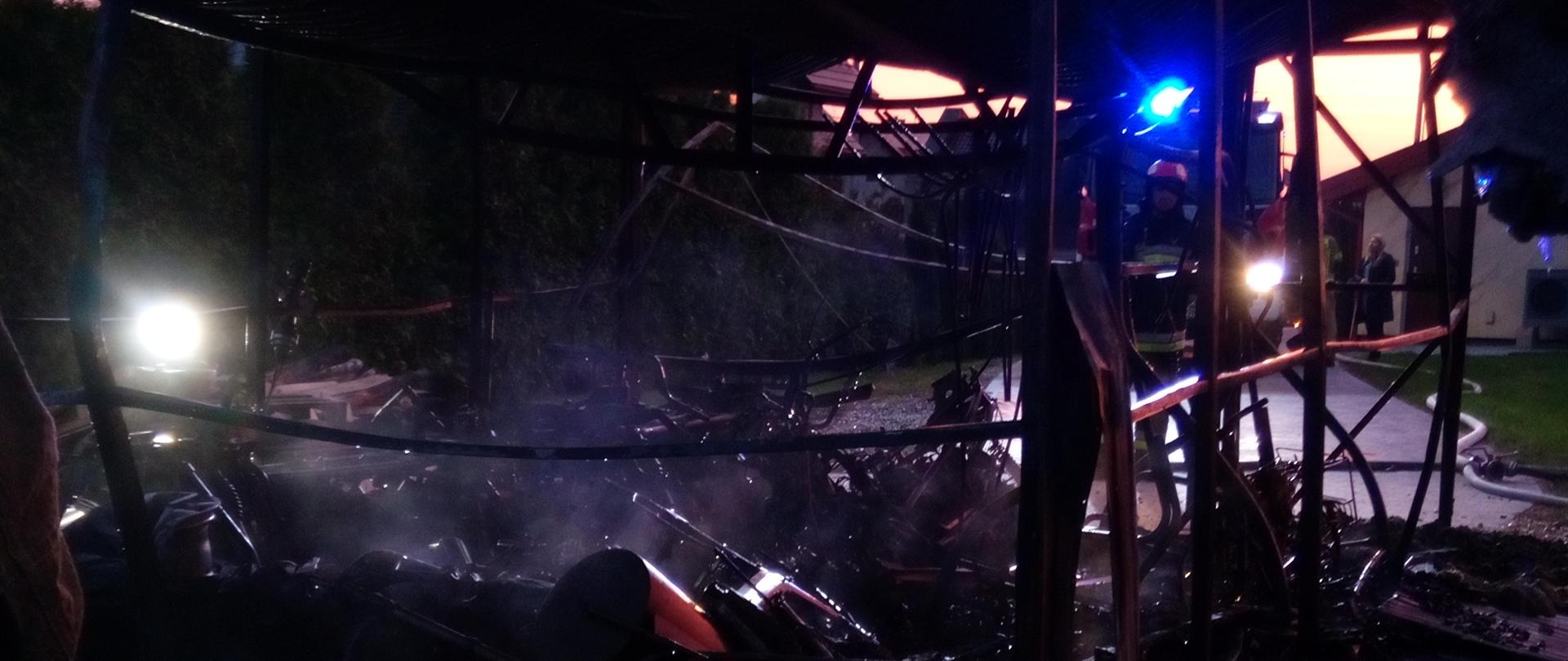 Zdjęcie przedstawia doszczętnie spalony garaż wolnostojący. Został sam "Szkielet" Na zewnątrz panuje półmrok. W oddali widać niebieską poświatę od sygnałów alarmowych samochodu PSP.