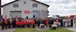Na zdjęciu widać dużą grupę osób stojącą na trawniku przed budynkiem straży pożarnej. Nad budynkiem powiewa polska flaga. Na pierwszym planie po lewej stronie stoją osoby trzymające sztandary. W centrum zdjęcia znajduje się pomnik w kształcie kamienia z tablicą i kwiatami. Większość osób jest ubrana odświętnie, w tym wiele dzieci w czerwonych koszulkach. W tle widoczne są wozy strażackie.