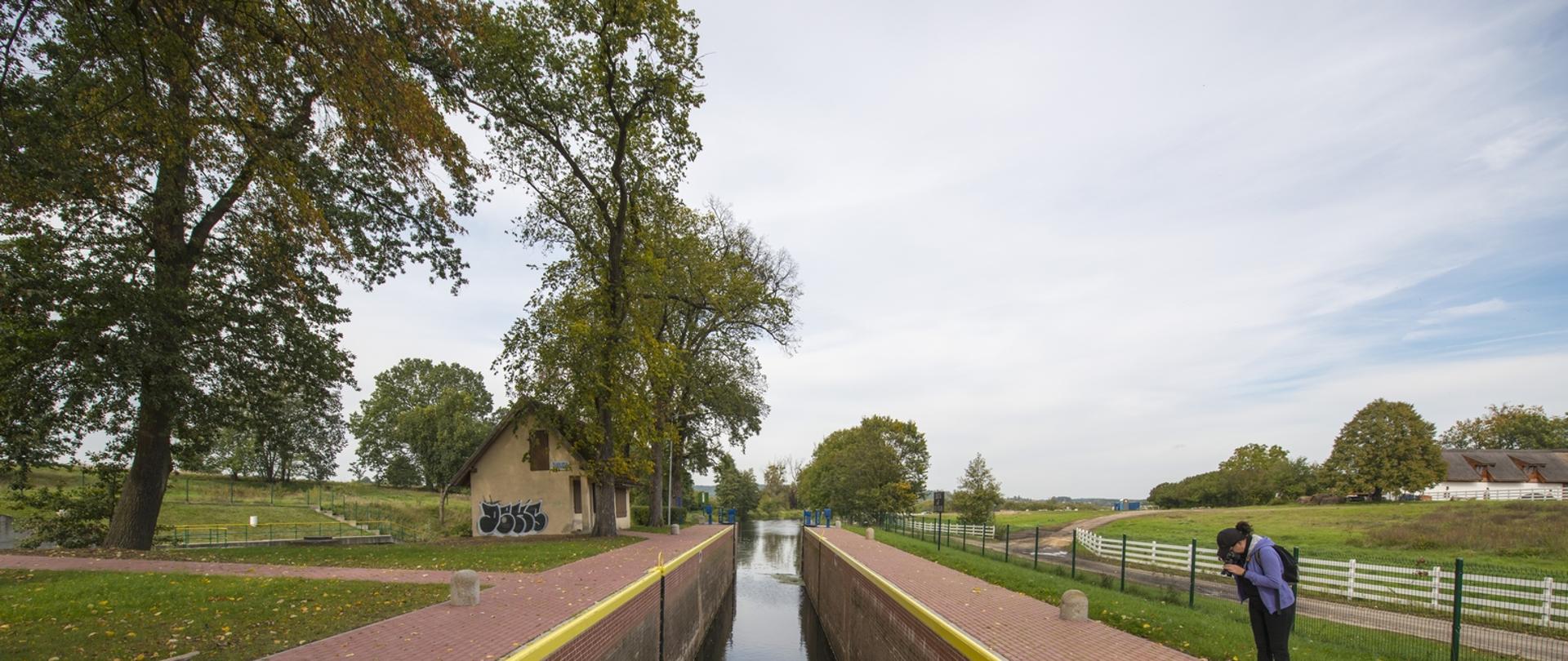 Śluza Nr 8 na Górnonoteckim odcinku drogi wodnej Warta – Kanał Bydgoski. Jednokomorowa o wymiarach komór 42,0x5,00 m wykonanych z betonu, cegły klinkierowej i ciosów kamiennych. Po prawej stronie jedna z osób - uczestników Pleneru Fotograficznego organizowanego przez tutejszy Regionalny Zarząd Gospodarki Wodnej w Bydgoszczy. Przed nami komora śluzy wypełniona wodą, w oddali zachmurzone niebo i po lewej stronie "śluzantówka". 