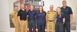 Wizyta strażaków z Attendorn. Salka edukacyjna w leszczyńskiej komendzie PSP. Na tle ściany, na której znajduje się wóz strażacki, do wspólnego zdjęcia pozują goście wraz z komendantem powiatowym, jego zastępcą oraz strażakiem z Leszna, który prowadził zajęcia edukacyjne.