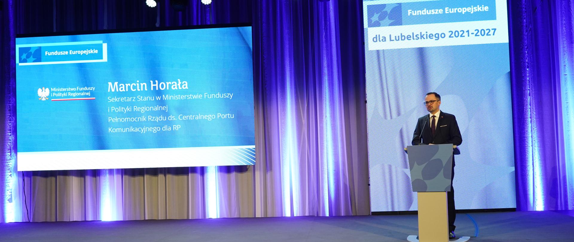 Wiceminister Marcin Horała stoi przy pulpicie na scenie i mówi do mikrofonu.