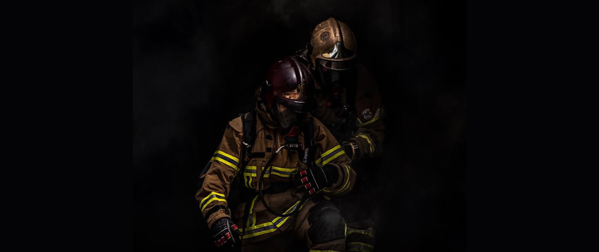Artystyczne zdjęcie dwóch strażaków na czarnym tle wspierających się i ukazujące ich poświecenie. Są ubraniu w umundurowanie specjalne wraz z aparatami oddechowymi oraz okopceni dymem.