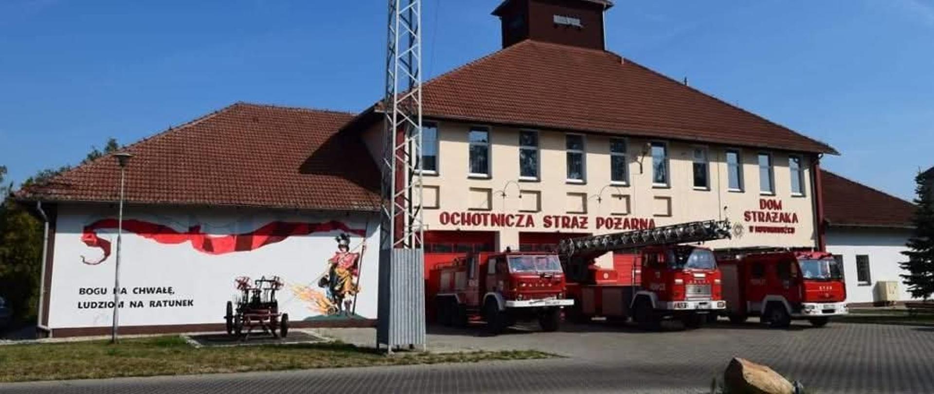 Szeroki plan zrobiony w świetle dziennym przedstawia budynek remizy Ochotniczej Straży Pożarnej w Nowogrodźcu. Jest to duży, dwupiętrowy budynek, pomalowany na jasny odcień écru, z dobudówką z lewej strony oraz dachem pokrytym ceglastą dachówką. Na środku dachu znajduje się mała wieża. Nad trzema bramami wjazdowymi wisi czerwony napis „OCHOTNICZA STRAŻ POŻARNA”. Nad wejściem do bocznego skrzydła widnieje mniejszy, ciemny napis „DOM STRAŻAKA w Nowogrodźcu”.
​Od strony wejścia ustawione są trzy czerwone wozy strażackie z drabinami. Z lewej strony fasady widnieje malowidło przedstawiające strażaka z tarczą i włócznią, oraz czerwoną, pofalowaną wstęgę. Poniżej w dwóch linijkach widnieje napis „BOGU NA CHWAŁĘ, LUDZIOM NA RATUNEK”. Przez budynkiem w centralnej części stoi duży, wysoki, metalowy maszt.
​Przed budynkiem z lewej strony stoi zabytkowy, pomalowany na ciemne odcienie wóz strażacki z zamontowanym na stałe działkiem wodnym. Na pierwszym planie widać trawę, zieleń i chodnik wyłożony kostką brukową. W oddali widać niebieskie niebo bez chmur.