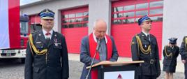 Zdjęcie przedstawia ks. Krzysztofa Kiełbowicza w trakcie przemówienia podczas uroczystości. Obok stojący Komendant Powiatowy PSP w Lwówku Śląskim st. bryg. Mariusz Mróz oraz dowódca uroczystości kpt. Bartosz Lisowski. 