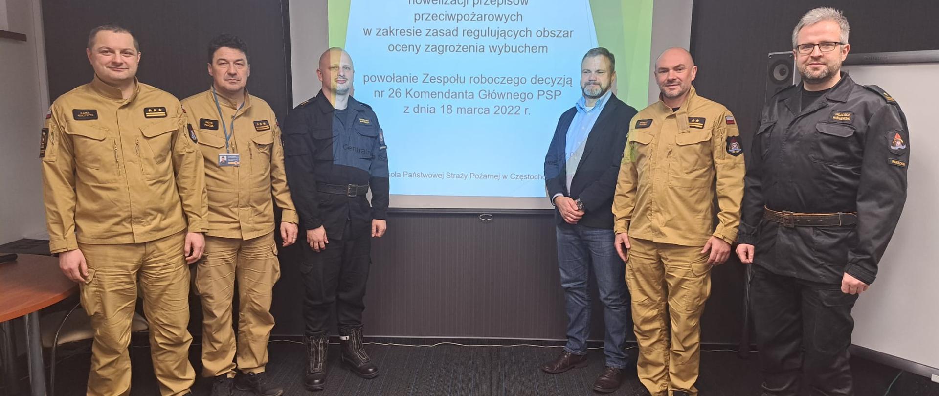Po lewej stronie stojący Komendant Centralnej Szkoły PSP w towarzystwie dwóch oficerów PSP, po prawej stronie stoi dwóch oficerów PSP oraz mężczyzna. W tle, na środku ekran multimedialny z wyświetlonym slajdem