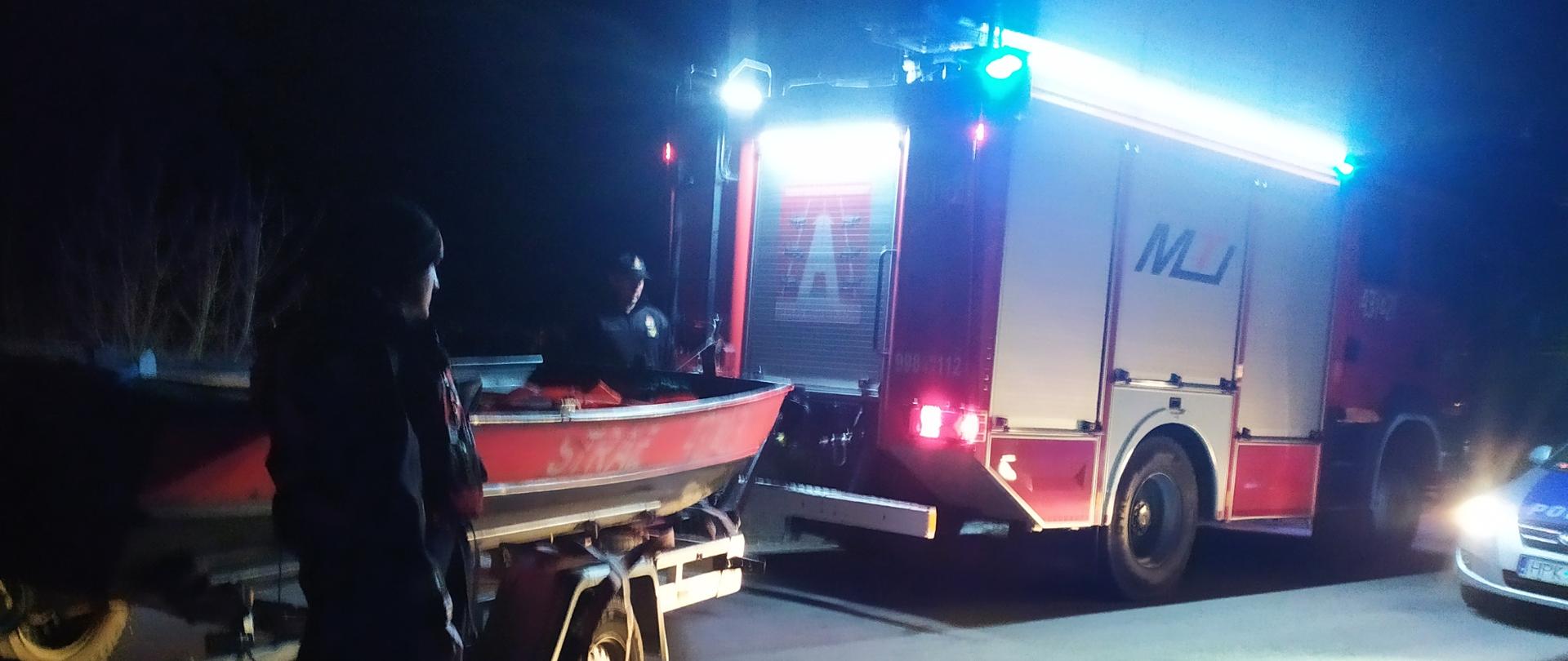 Na zdjęciu, zrobiony w porze nocnej widać wóz strażacki z podpiętą łodzią płaskodenną, a także radiowóz policyjny. Przed i za łodzią widać dwóch strażaków Państwowej Straży Pożarnej, ubranych w kombinezony służące działaniom ratowniczym na wodzie.