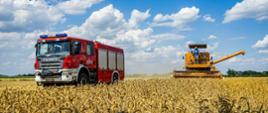 Duży czerwony samochód pożarniczy a za min żółty kombajn stoją w zbożu, nad nimi błękitne niebo.