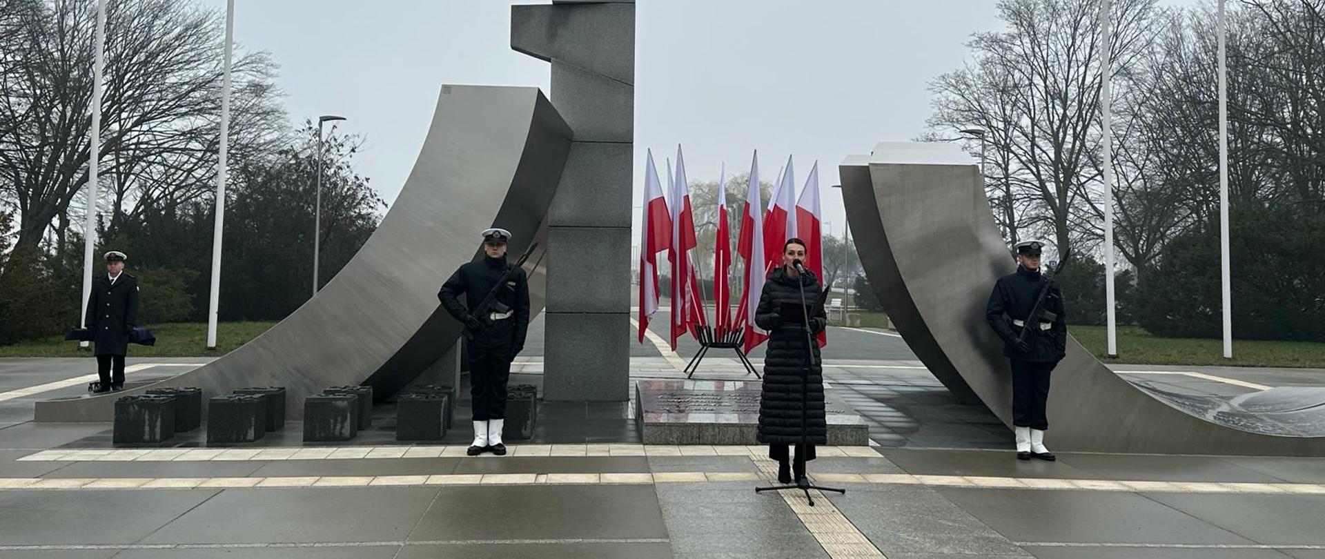 pomnik, przed nim kobieta (wojewoda) wygłaszająca przemówienie, obok dwóch marynarzy i flagi Polski
