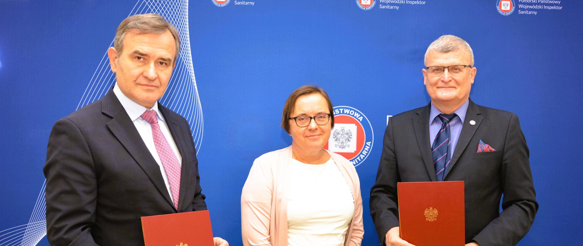 Na tle ścianki z logotypem Państwowej Inspekcji Sanitarnej stoją trzy osoby. Po bokach stoi dwóch mężczyzn, w środku kobieta. Mężczyźni trzymają w dłoniach teczki z podpisanymi dokumentami.