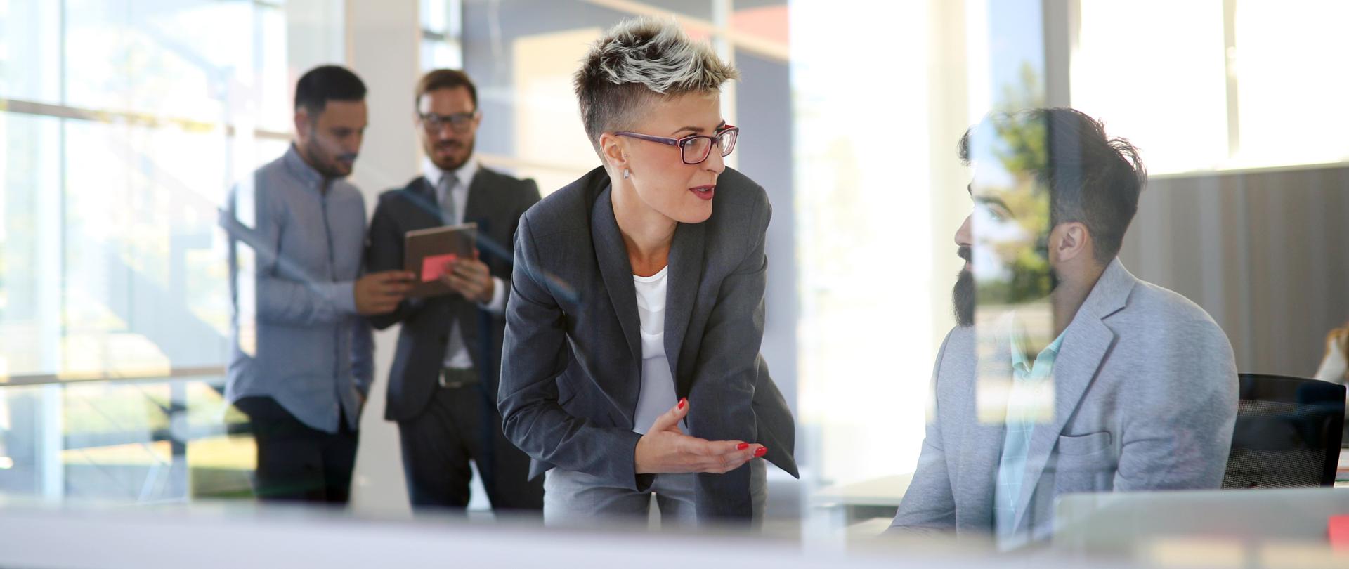 Pracownicy w biurze. Na pierwszym planie dyskutują siedzący mężczyzna ze stojącą obok kobietą.