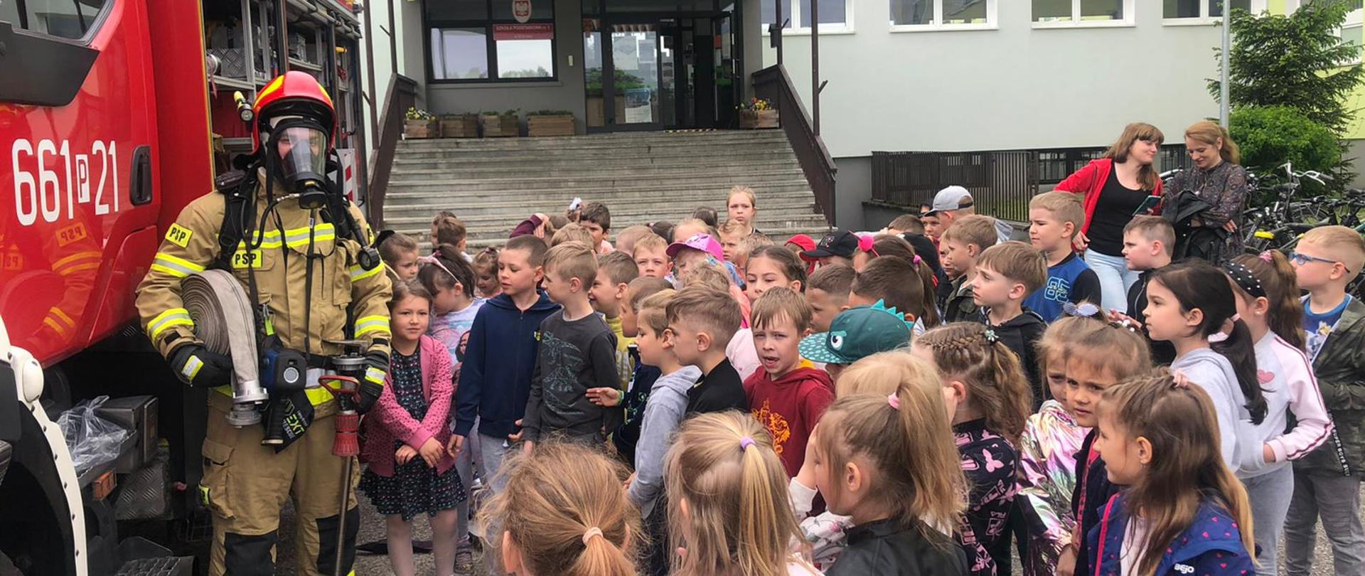 Strażak prezentuje dzieciom pełne wyposażenie strażaka do gaszenia pożarów.