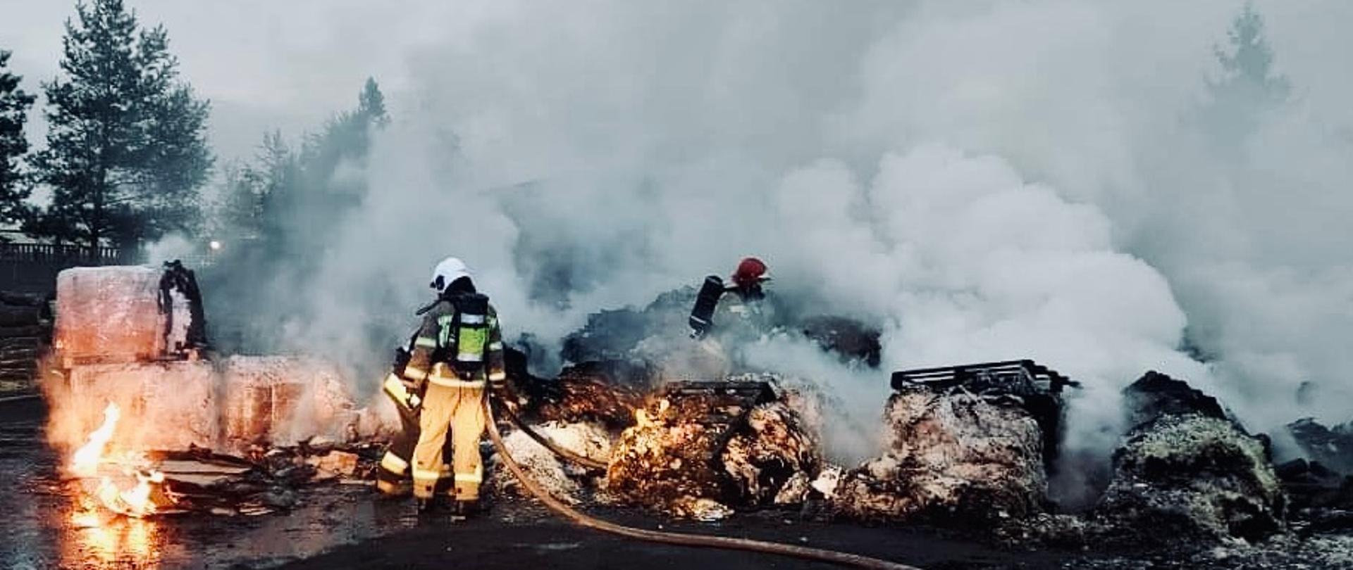 Na zdjęciu widać strażaka OSP oraz strażaka PSP w pełnych ubraniach ochronnych którzy podają prądy wody na palące się palety ze sprasowaną makulaturą.