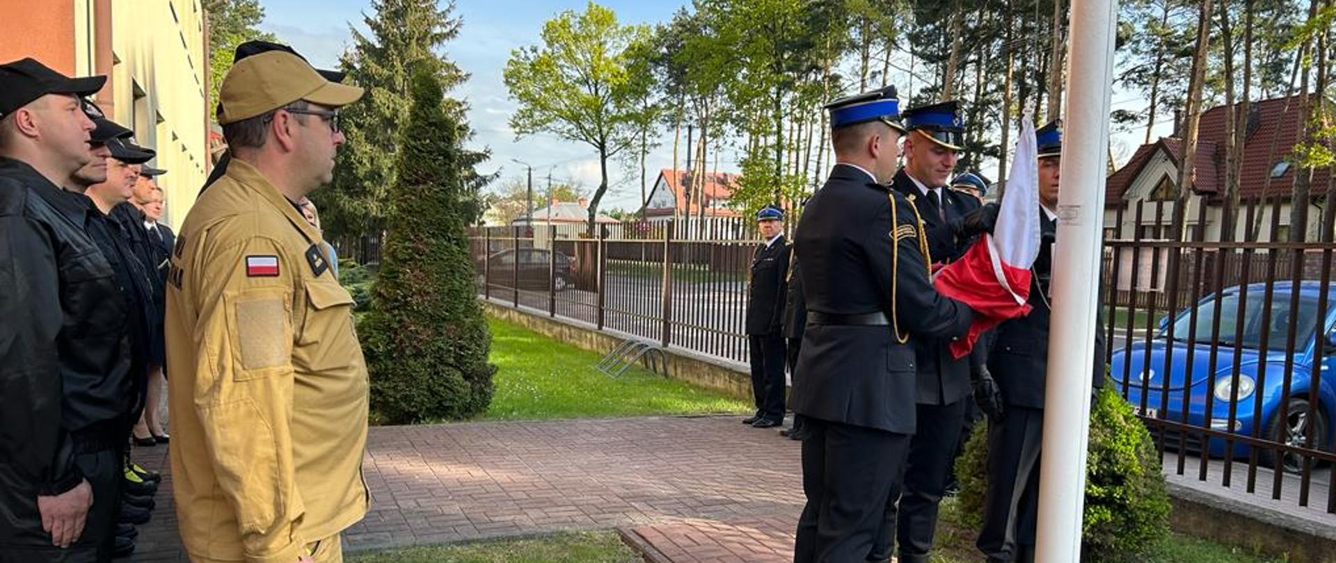 Zdjęcie przedstawia strażaków zgromadzonych przy maszcie na którym podniesiona została flaga państwowa. Strażacy ubrani są w umundurowanie koszarowe. Na pierwszym planie widać poczet flagowy podnoszący flagę do góry. Natomiast w tle widoczny jest Komendant Powiatowy w mundurze wyjściowym. W tle widać wysokie iglaste drzewa.