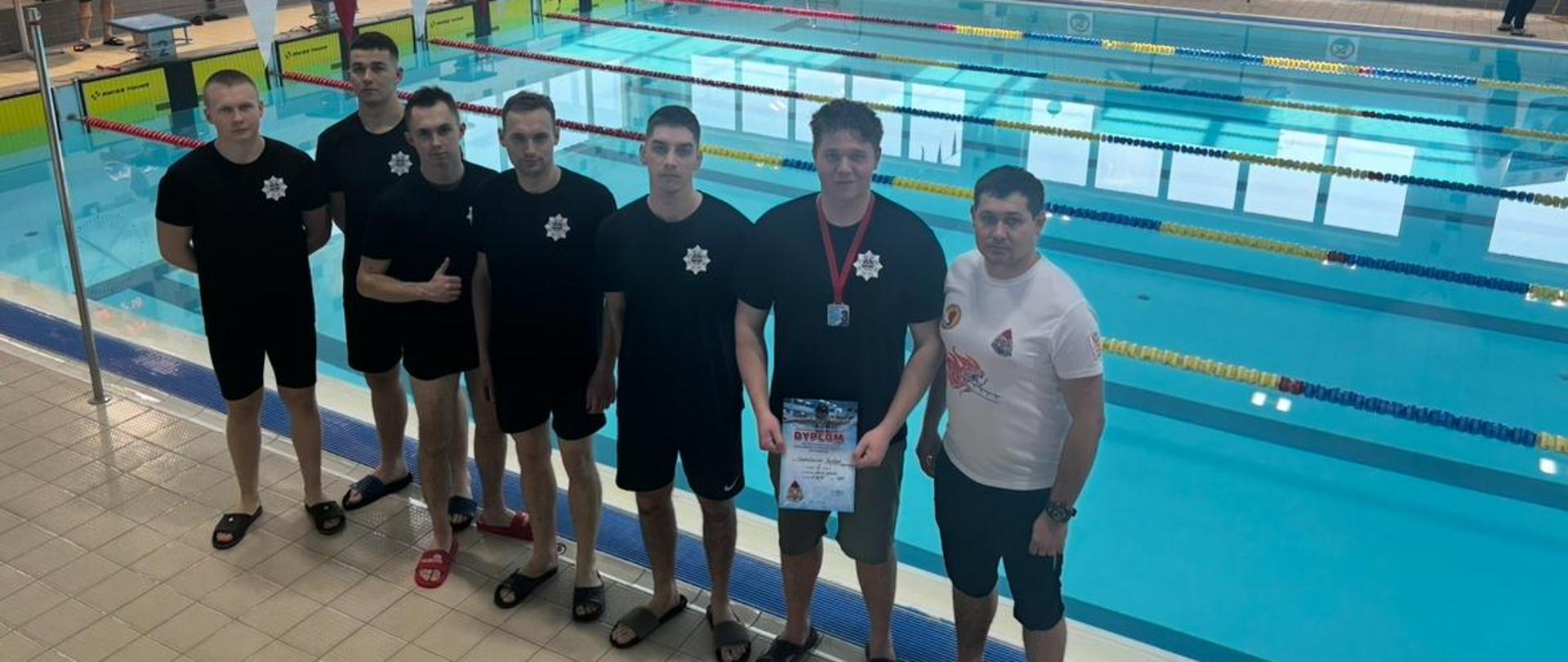 Pamiątkowe zdjęcie zawodników reprezentujących Centralną Szkolę PSP na Mistrzostwach Polski Służb Mundurowych w Pływaniu (6 osób) wraz z opiekunem na tle basenu