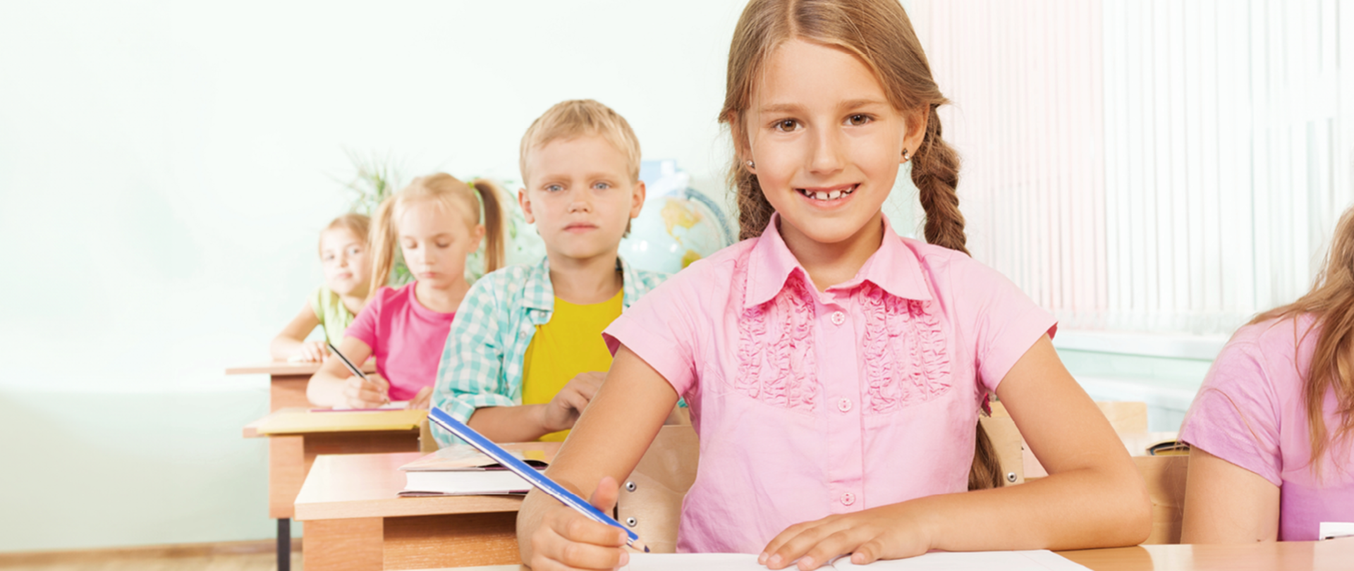 Dzieci siedzące przy ławkach w klasie. Na pierwszym planie uśmiechnięta dziewczynka z kredką w dłoni. 
