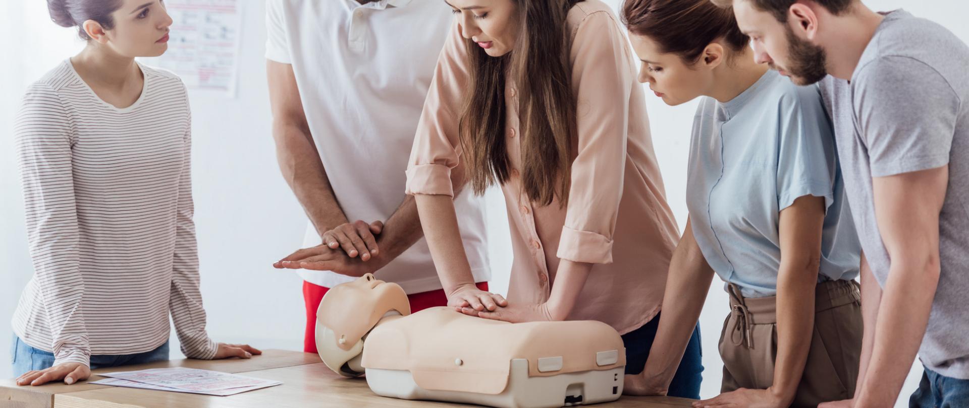 Na zdjęciu jest widoczny fantom leżący na stole, uciskany przez kobietę. Obok znajdują się osoby postronne tj. dwie kobiety i dwóch mężczyzn.