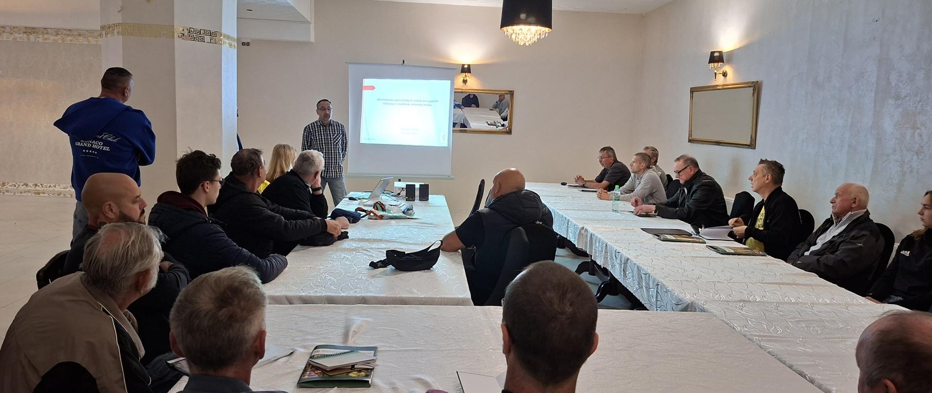 Sala konferencyjna z uczestnikami siedzącymi przy stołach, w tle inspektor w trakcie swojego wystąpienia i wyświetlona prezentacja
