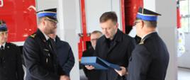 6. W garażu Komendy Powiatowej Państwowej Straży Pożarnej w Świeciu po lewej stronie stoi bokiem Komendant Powiatowy Państwowej Straży Pożarnej w Świeciu starszy brygadier Piotr Tkaczyński, który wręczył pamiątkową statuetkę Panu Ministrowi Łukaszowi Schreiber. Obok Pana Ministra stoi lewym profilem Kujawsko-Pomorski Komendant Wojewódzki Państwowej Straży Pożarnej starszy brygadier Jacek Kaczmarek. W tle stoją pozostali uczestniczy spotkania a za nimi widoczny bok samochodu strażackiego.
