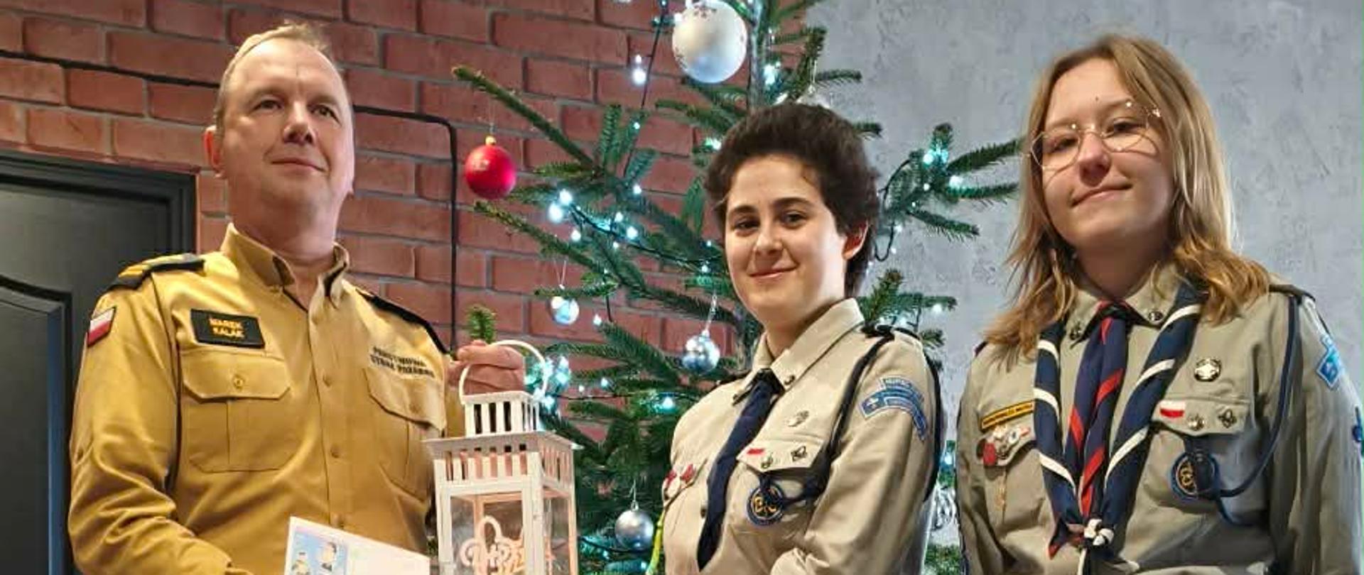 Zdjęcie przedstawia komendanta w mundurze piaskowym odbierającego światełko pokoju od harcerzy na tle choinki