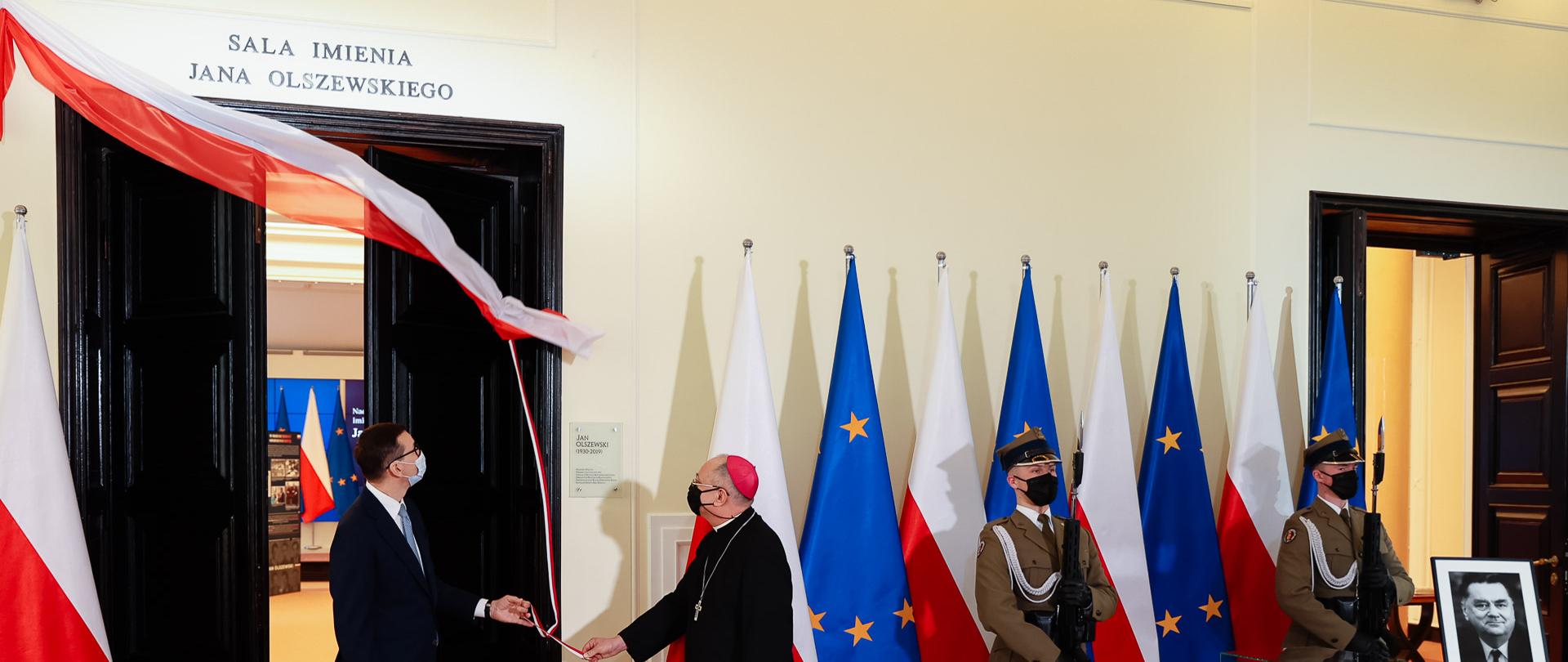 Premier oraz biskup odsłaniają napis nad salą, obok stoją żołnierze.