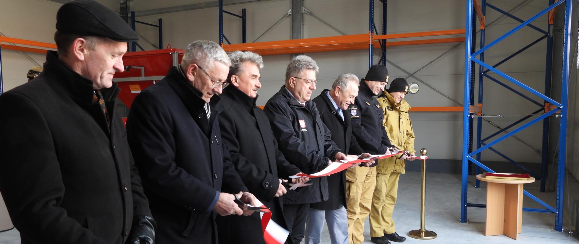 Wnętrze hali magazynowej podczas uroczystego otwarcia obiektu. Kilku mężczyzn ubranych w ciemne płaszcze oraz dwóch strażaków w mundurach stoi w rzędzie i przecina biało-czerwoną wstęgę nożyczkami. Obok znajduje się złoty słupek z czerwonym sznurem, a w tle widoczne są metalowe regały magazynowe oraz elementy wyposażenia hali.