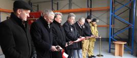 Wnętrze hali magazynowej podczas uroczystego otwarcia obiektu. Kilku mężczyzn ubranych w ciemne płaszcze oraz dwóch strażaków w mundurach stoi w rzędzie i przecina biało-czerwoną wstęgę nożyczkami. Obok znajduje się złoty słupek z czerwonym sznurem, a w tle widoczne są metalowe regały magazynowe oraz elementy wyposażenia hali.