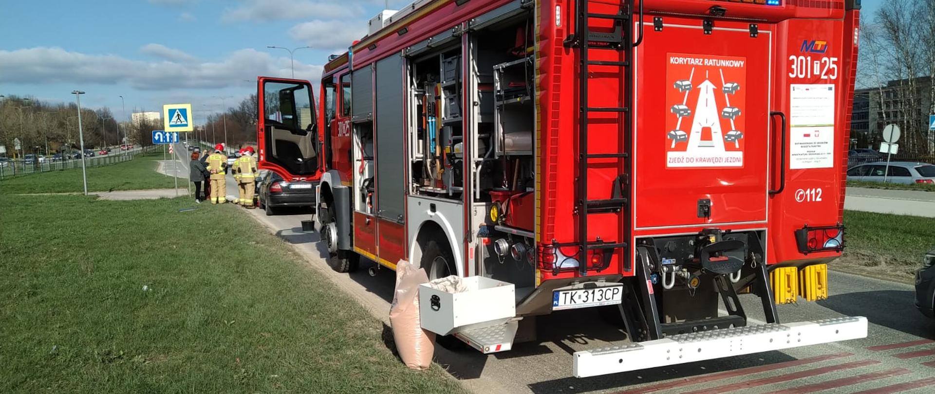 Zdjęcie przedstawia ulicę Solidarności. Na pierwszym planie stoi samochód pożarniczy, a przy jego kole znajduje się worek z sorbentem. W głębi widać grupę strażaków stojących przy uszkodzonych samochodach biorących udział w kolizji.