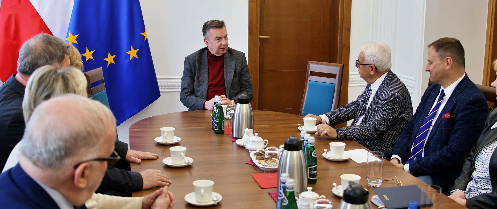 Przy owalnym stole siedzi kilka osób, u szczytu stołu minister Wieczorek, za nim pod ścianą flagi Polski i UE.