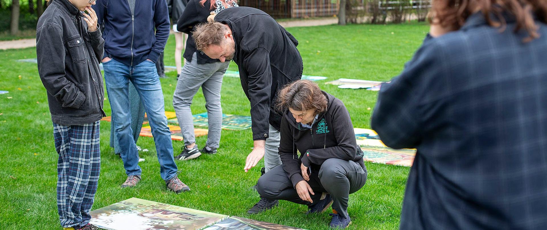 To zdjęcie przedstawia grupę ludzi, która stoi na trawiastym terenie, prawdopodobnie w parku lub ogrodzie. Wszyscy są odwróceni w stronę kamer. Na ziemi, tuż przed nimi, leżą na trawie obrazy, które prawdopodobnie są ich dziełami. Ludzie oglądają je i dyskutują, niektórzy pochylają się, aby lepiej im się przyjrzeć.