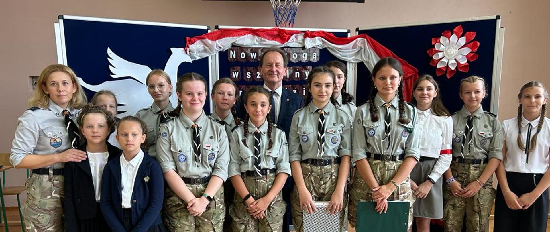 Pod ścianą sali gimnastycznej stoi wiceminister Bernacki, przed nim szereg ubranych w mundury dzieci.