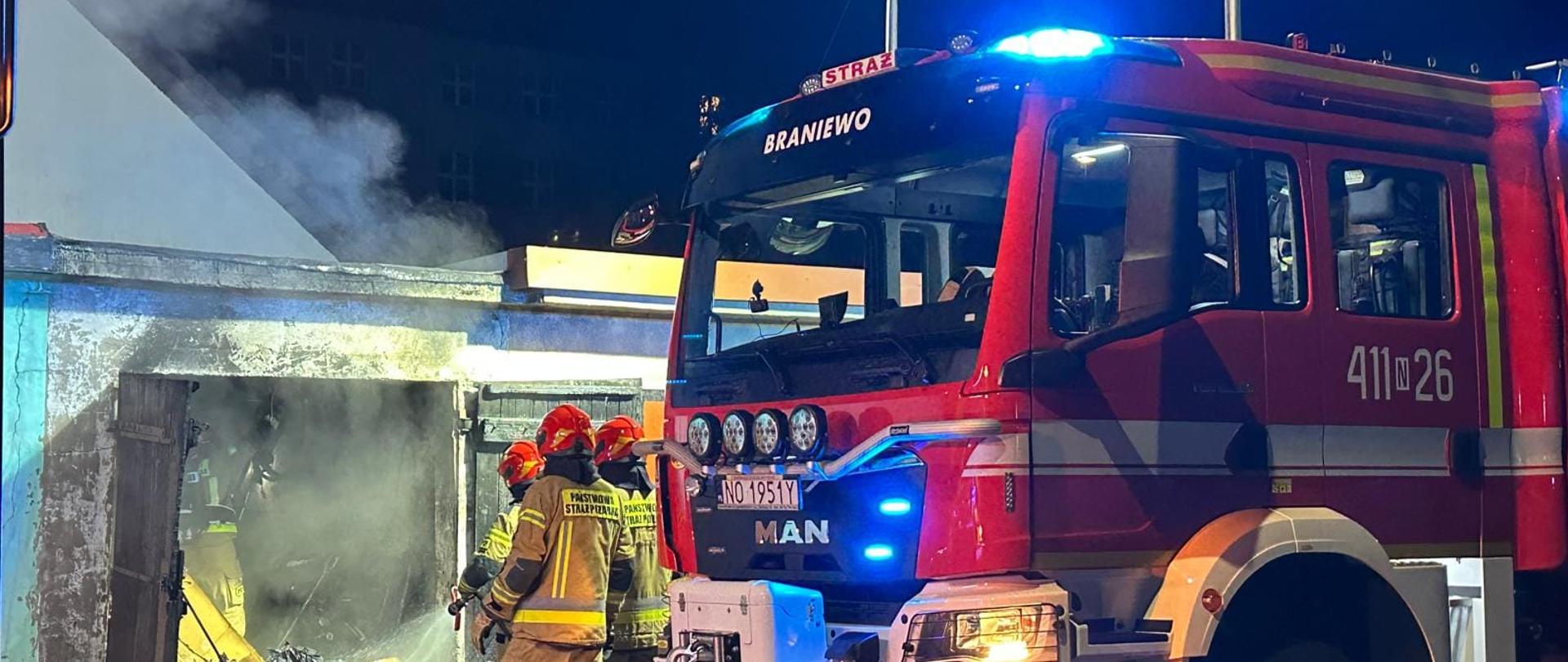 Z garażu w którym był pożar wydobywa się dym. Z prawej strony duży samochód strażacki i strażacy.