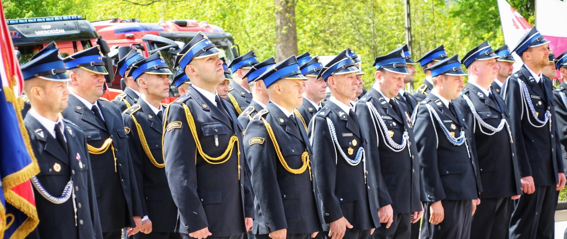 Strażacy w granatowych mundurach wyjściowych stoją w szeregu przed remizą OSP Mieroszów