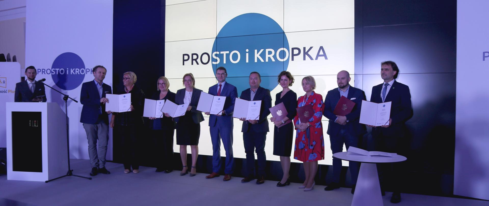 Jedenaście osób na scenie, dziewięć osób trzyma teczki z dokumentami, za nimi na ekranie hasło 'Prosto i kropka", po lewej stronie sceny stoi mównica, po prawej stronie stolik.