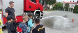 Dziecko leje wodę z prądownicy strażackiej, celuje w pachołek, pomaga mu strażak. 